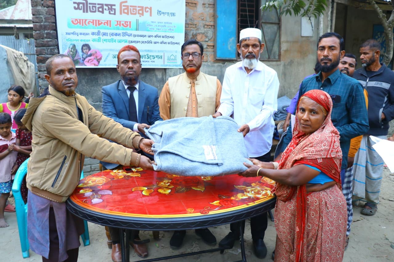 খোলা উঠোনে লাল টেবিলের ওপর দিয়ে একজন নারীর হাতে ধূসর কম্বল দেওয়া হচ্ছে।